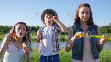 孩子们玩得很开心，情绪激动的姐<strong>妹</strong>和戴帽子的小男孩在湖边的草地上对着蓝天吹<strong>肥</strong>皂泡